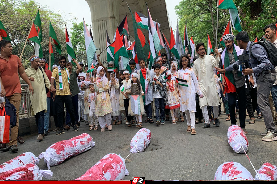প্রতীকী রক্তমাখা পোশাক, ব্যান্ডেজ, ফিলিস্তিনের পতাকা হাতে  প্রতীকী লাশের সামনে মিছিন করছে শিশুরা, তাদের সঙ্গে মিছিলে অংশ নেয় নানা বয়সের মানুষ